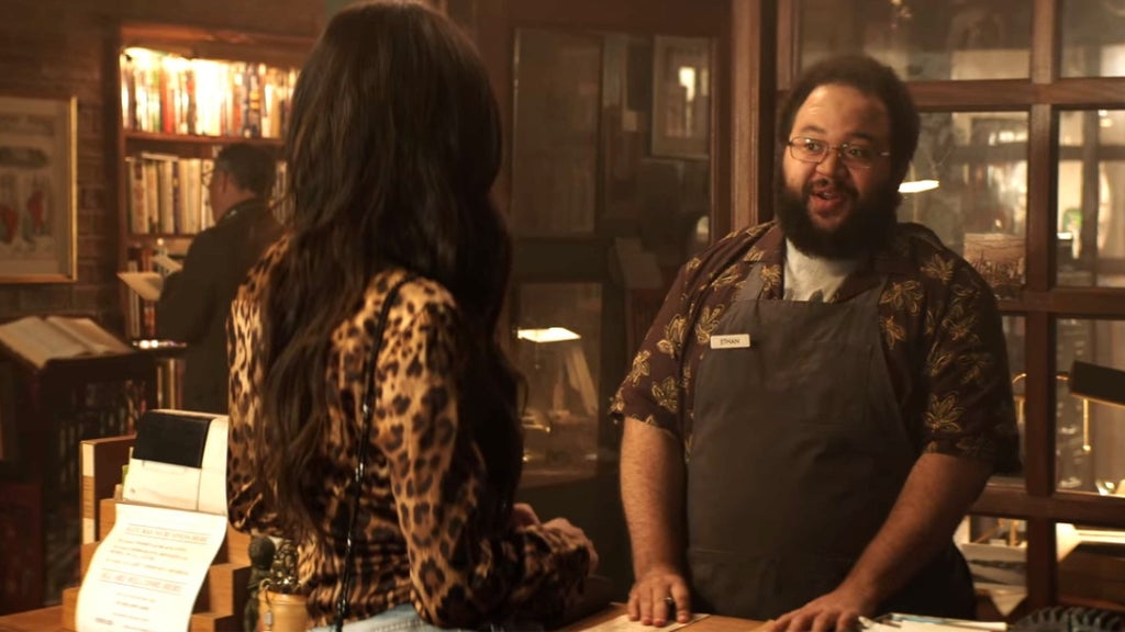 Inside a warmly lit bookstore, a cheerful man wearing glasses, a floral button-up shirt, and a dark apron with a name tag reading “Ethan” stands behind the counter speaking animatedly with a customer. The woman, seen from behind, has long wavy hair and wears a leopard-print top. The cozy atmosphere is filled with bookshelves, wooden furniture, and soft lighting, suggesting a friendly and inviting setting.
