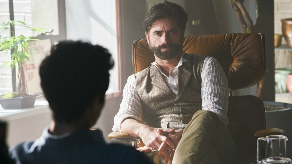 A man with dark hair and a full beard sits confidently in a mid-century modern leather chair, wearing a beige plaid vest over a white shirt with rolled sleeves. He faces another person who is partially visible in the foreground, suggesting a conversation or therapy session. The setting is softly lit by natural light from a nearby window, creating a calm, introspective atmosphere in the room decorated with plants and books.