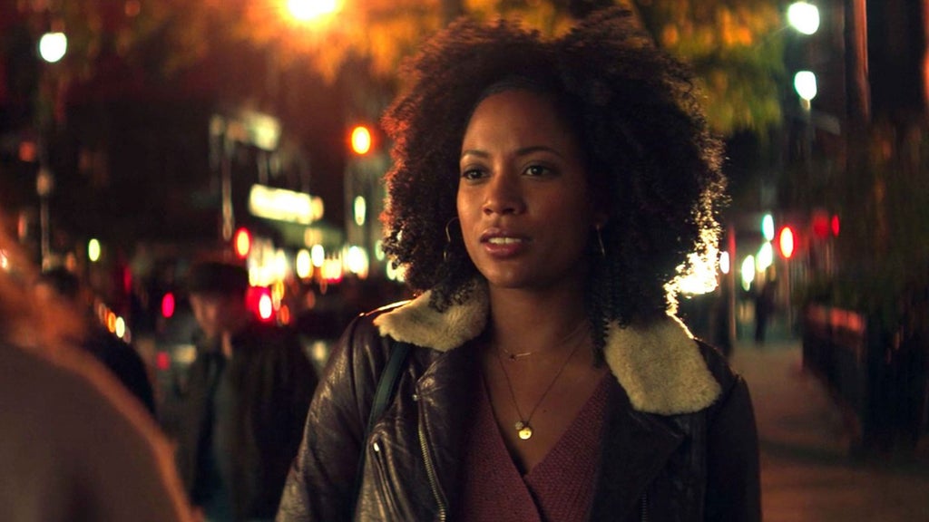 A woman with natural curly hair walks through a brightly lit city street at night, framed by the warm glow of streetlights and storefronts. She wears a leather jacket with a shearling collar over a maroon top, accessorized with layered gold necklaces. Her expression is composed but thoughtful, as if she's in the middle of an important conversation or decision. The blurred background looks like a city with streetlights and lit signs