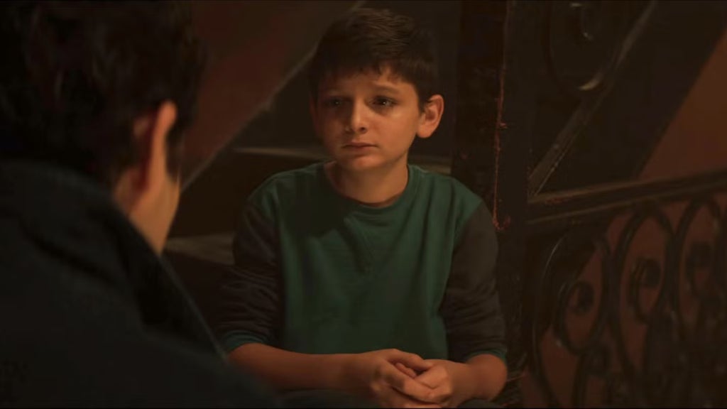 A young boy sits on a staircase in a dimly lit building, looking up with a solemn and slightly teary-eyed expression. He wears a green long-sleeve shirt and clasps his hands together in front of him. Facing him is a man seen from behind, suggesting a serious or emotional conversation is taking place. The atmosphere feels intimate and tense, highlighting the boy's vulnerability.