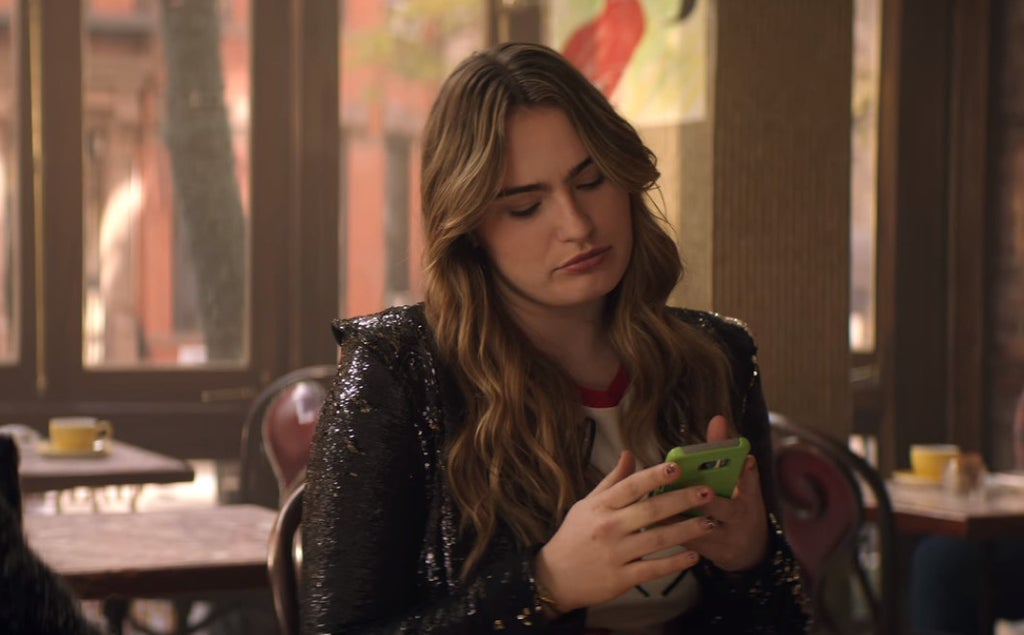 A young woman with long, wavy hair sits at a table in a cozy café, wearing a sparkly black jacket over a graphic tee. She looks down at her bright green phone with a disinterested or mildly annoyed expression, her fingers poised over the screen. Sunlight streams in through large windows behind her, casting a warm, natural glow over the relaxed, daytime setting. Yellow coffee cups are visible on nearby tables, adding a casual, social atmosphere.