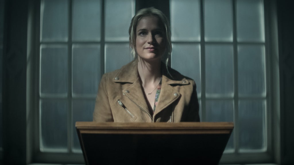 A woman with blonde hair pulled back stands behind a wooden podium in a dimly lit room, softly illuminated by light streaming through large, foggy-paned windows behind her. She wears a tan suede jacket and has a composed yet mysterious expression, with shadows casting a dramatic ambiance around her.