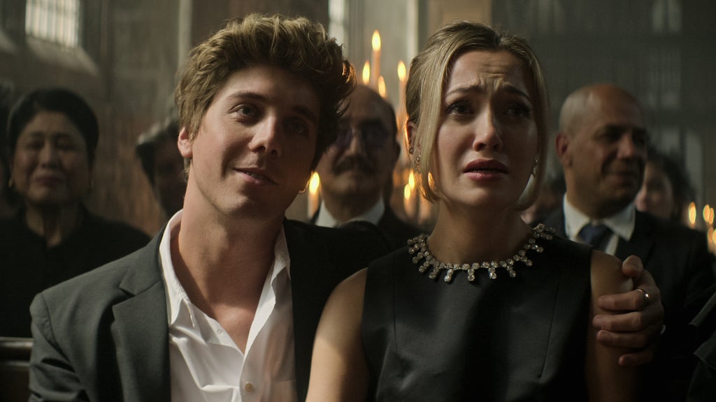 A well-dressed young couple sits in a candlelit church pew during a somber ceremony. The man, wearing an open-collar suit, looks relaxed and slightly amused, while the woman beside him appears emotional, eyes glistening with tears and lips trembling as she tries to hold back sobs.