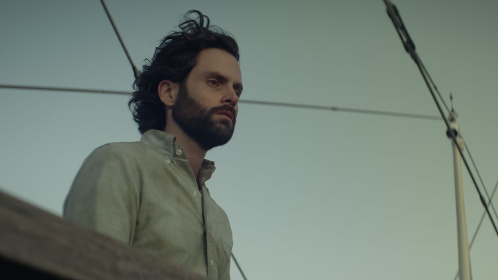 A man with dark, wavy hair and a full beard stands on a boat, gazing into the distance with a somber, contemplative expression. He wears a light-colored button-up shirt, partially unbuttoned, as the soft lighting of dusk or dawn casts a moody glow. Overhead, rigging lines and parts of the mast hint at the nautical setting, adding to the introspective atmosphere.