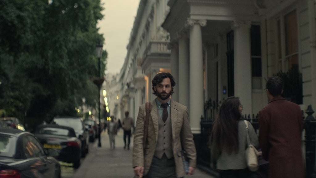 A man with curly hair and a beard, dressed in a brown checkered blazer, vest, and tie, walks alone down a dimly lit, upscale residential London street lined with white townhouses and parked cars. He carries a brown shoulder bag and a book, looking serious and contemplative. Other pedestrians walk in the opposite direction, and the atmosphere is moody and overcast.