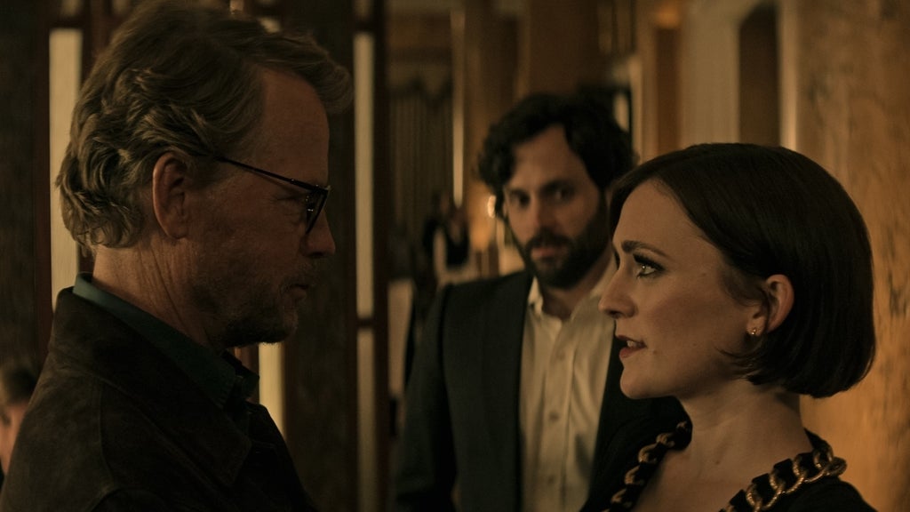 In a dimly lit, upscale interior, a tense moment unfolds as a woman with short brown hair and a chunky gold chain necklace stares down an older man with glasses and a beard. Behind them, a man with curly hair and a serious expression looks on, partially obscured by shadows. The atmosphere is heavy with unspoken conflict.