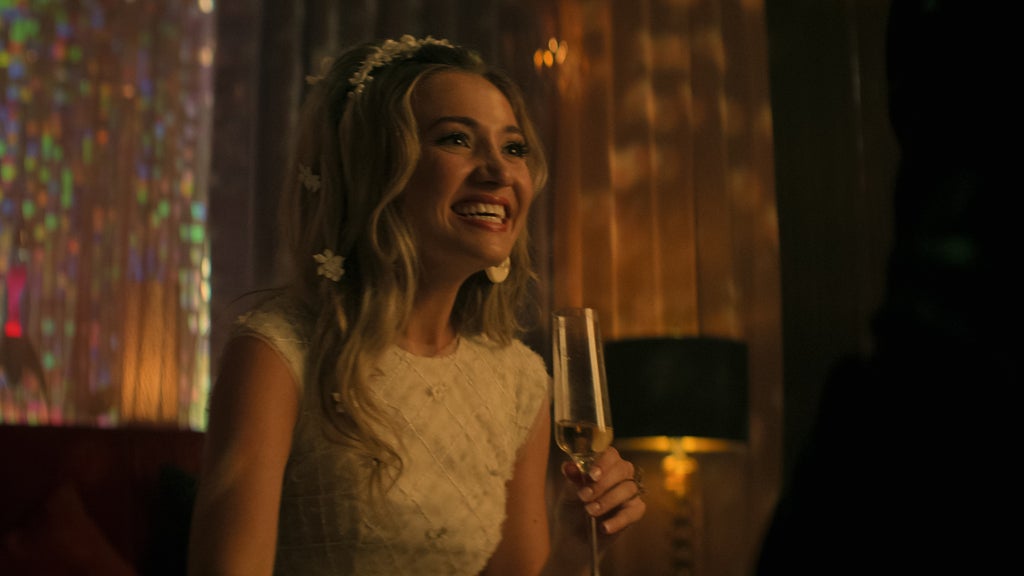 A smiling blonde woman in a white dress sits in a warmly lit, festive room holding a champagne flute. Her long hair is styled with white flower accessories, giving her a whimsical and celebratory appearance. Behind her, colorful lights and draped curtains create a dreamy, party-like ambiance. Her expression is joyful, but her smile is too big and forced, suggesting she's in mental distress