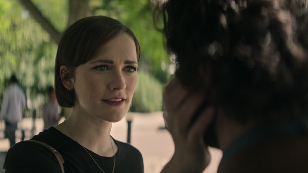A woman with short brown hair gently touches the face of a man whose back is to the camera. They are standing outdoors under the shade of green trees, with soft sunlight filtering through the leaves. The scene conveys an intimate moment between them.
