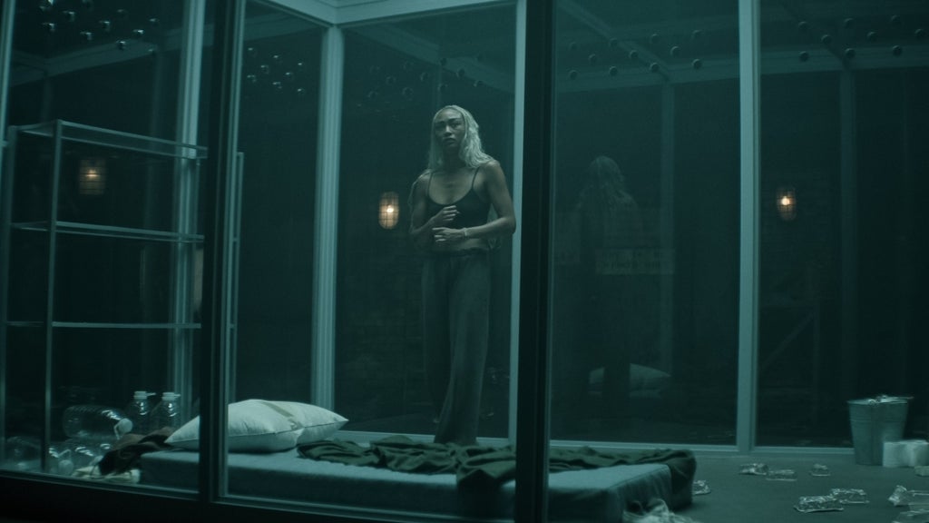 A woman with long, platinum blonde hair stands inside a dimly lit, glass-walled room that resembles a high-security cage or holding cell. She wears a black tank top and dark pants, appearing tense and cautious as she clutches her stomach. The sparse room contains a thin mattress on the floor with a pillow and blanket, empty bottles, and scattered trash, reinforcing the sense of confinement and isolation.