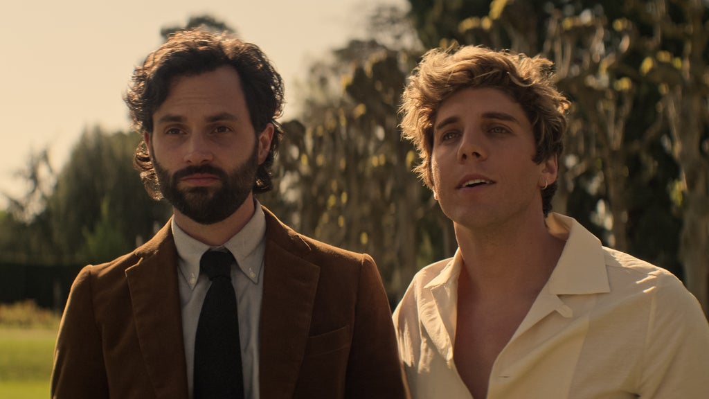 A man in a suit with dark curly hair stands beside a casually dressed blonde man outdoors in golden afternoon light. Joe wears a brown corduroy blazer and dark tie, looking serious and contemplative, while the man beside him smiles slightly, gazing into the distance with a relaxed demeanor. Behind them, tall trees blur in the warm background.