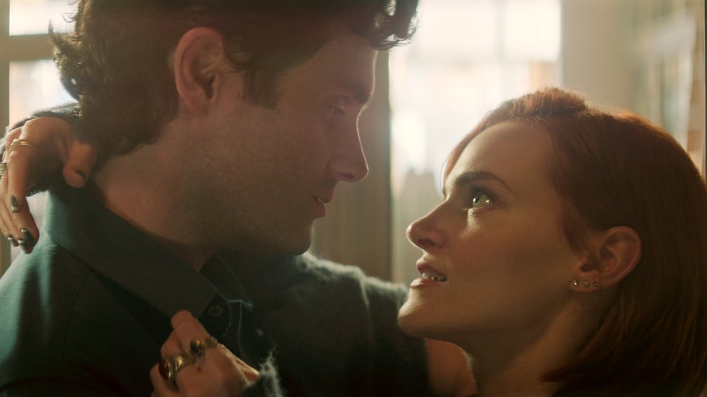 A man and a woman share an intense, intimate moment, holding each other closely and gazing into each other's eyes. The woman's arms are wrapped around the man's neck, her fingers adorned with rings, while the soft sunlight filtering through the background gives the scene a dreamy, romantic glow. Their expressions are filled with emotion, suggesting both connection and complexity in their relationship.