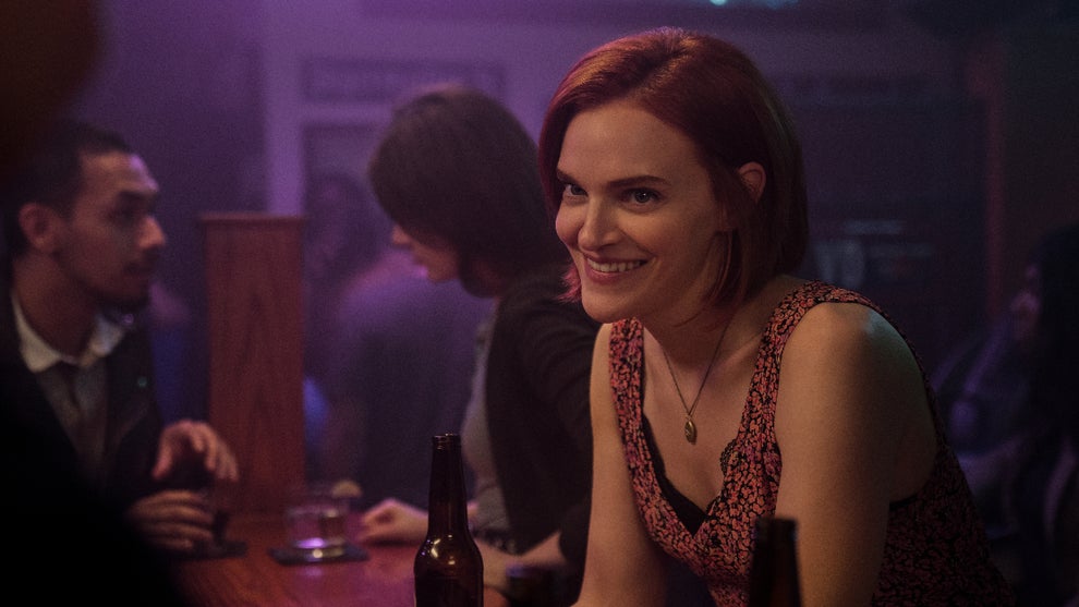 A woman with short red hair and a mischievous smile sits at a bar in a dimly lit, neon-hued setting. She wears a pink and black patterned top and a gold pendant necklace, leaning forward slightly as she engages in conversation. A bottle sits on the counter in front of her, and the background is filled with the blurred figures of other patrons, giving the scene a casual, lively nightlife vibe.