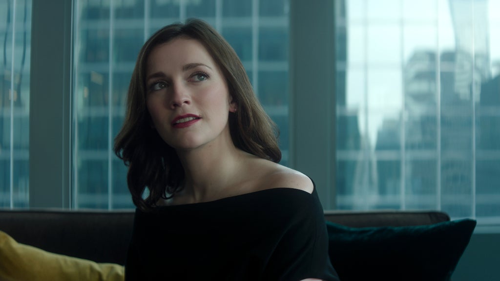 A woman with wavy brown hair and a poised expression sits on a couch in a modern high-rise apartment, framed by large windows with a cityscape in the background. She wears an elegant off-the-shoulder black top and gazes off to the side with a tense expression, as daylight streams in behind her, casting a cool, polished ambiance.