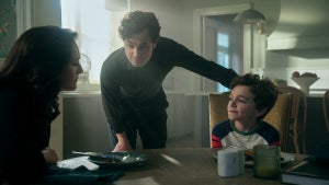 A man dressed in black leans over a dining table, placing a reassuring hand behind a young boy who sits smiling in a high-backed chair. The boy, wearing a colorful striped shirt, looks up at him with affection. Across the table, a woman with dark hair sits in soft focus, her expression contemplative. Morning light filters through the window, casting a warm, domestic glow over the cozy kitchen setting.
