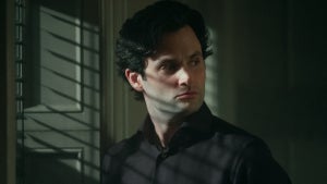 A man with wavy dark hair and a serious expression stands partially in shadow, looking over his shoulder with a wary, alert gaze. He wears a dark collared shirt, and light from nearby window blinds casts dramatic lines across his face and chest. The atmosphere is tense and suspenseful, suggesting a moment of secrecy or danger.