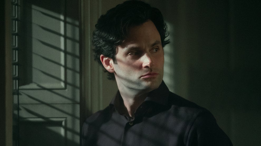 A man with wavy dark hair and a serious expression stands partially in shadow, looking over his shoulder with a wary, alert gaze. He wears a dark collared shirt, and light from nearby window blinds casts dramatic lines across his face and chest. The atmosphere is tense and suspenseful, suggesting a moment of secrecy or danger.