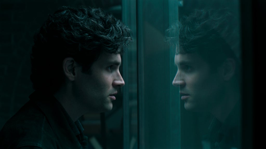 A man with dark, curly hair stares intently at his own reflection through a glass wall in a dimly lit, teal-toned space. The mood is tense and introspective, highlighting an internal struggle or moment of deep contemplation