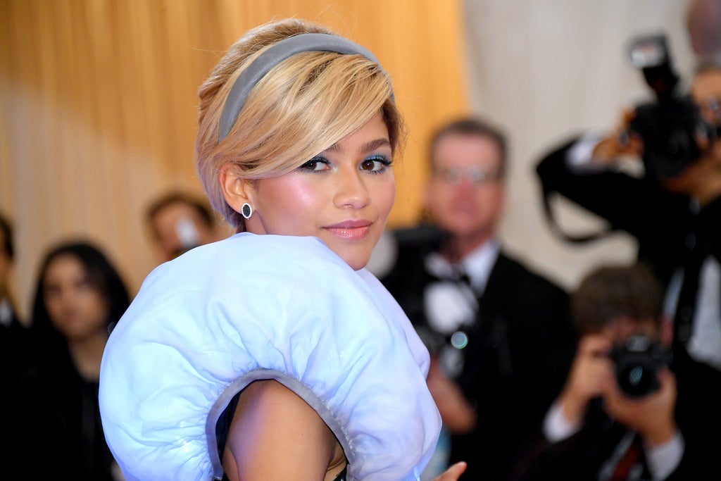 Zendaya channels Cinderella with a soft blonde updo and silver headband, wearing a voluminous blue ruffled detail on her shoulder. Her dewy pink makeup and pastel tones give a modern fairytale vibe on the Met Gala carpet.