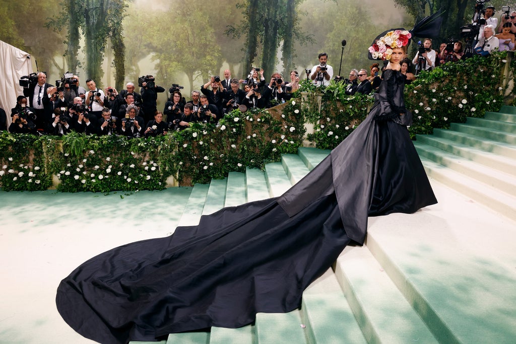 Zendaya stands atop the Met Gala steps in a sweeping black off-the-shoulder gown with an exaggerated train cascading down the staircase. She wears an oversized headpiece made of vibrant faux roses and structured black mesh, as photographers capture the moment in a lush, garden-themed setting.