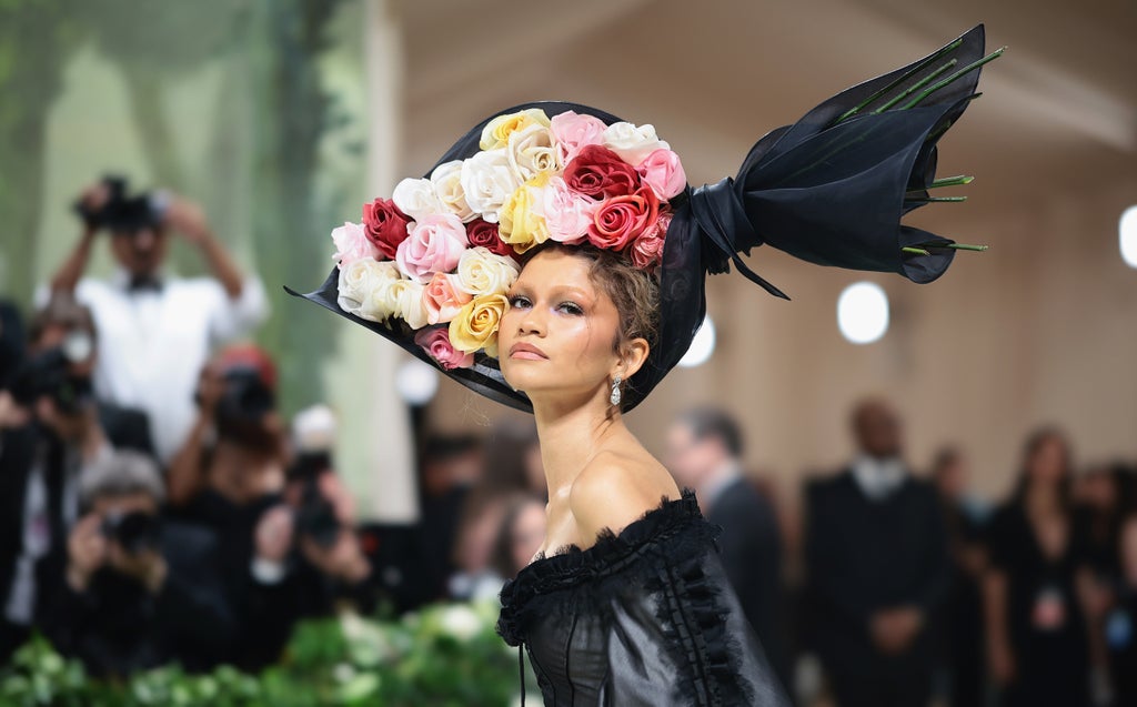 A close-up of Zendaya at the Met Gala wearing a dramatic floral headpiece made of large multicolored roses and black mesh. Her hair is styled in soft waves, and she wears sparkling drop earrings and a structured black gown with corset detailing and a romantic off-the-shoulder neckline.