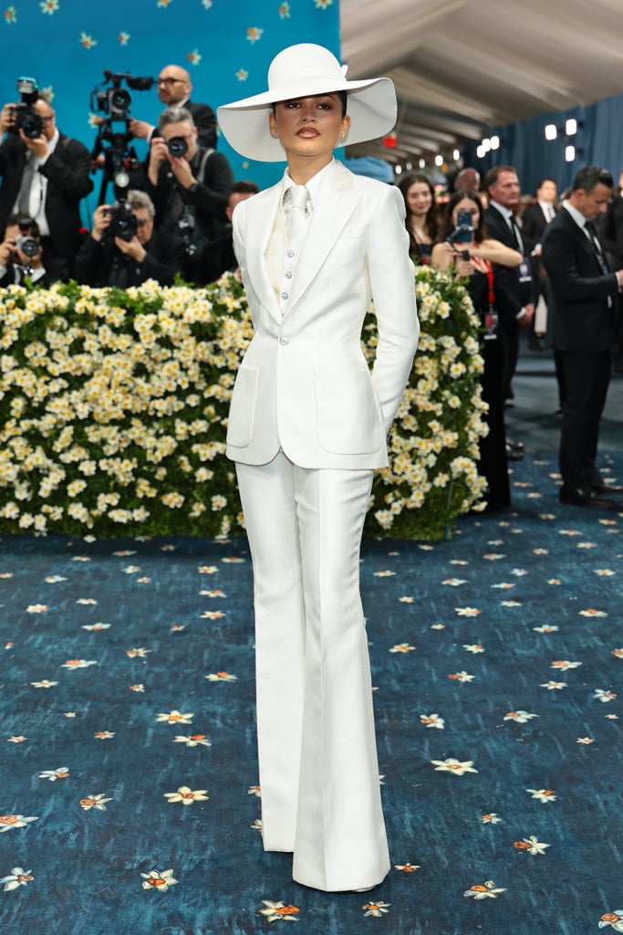 Zendaya stands confidently on the Met Gala red carpet wearing a sleek, all-white tailored suit with flared pants and a wide-brimmed hat. Behind her, photographers and onlookers line a backdrop of a flower-covered wall.