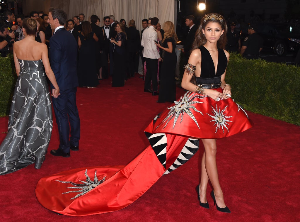 Zendaya poses on the Met Gala red carpet in a dramatic high-low red gown with embroidered sunburst designs, a black velvet bodice, and a structured train with graphic black-and-white lining. She accessorizes with a gold crown and bold sun-themed jewelry