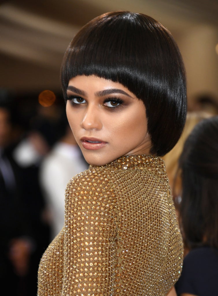 Zendaya gazes intensely over her shoulder in a gold, jewel-encrusted dress with a sculptural silhouette. Her sleek black bob with razor-straight bangs and dramatic eye makeup add a futuristic, high-fashion edge.