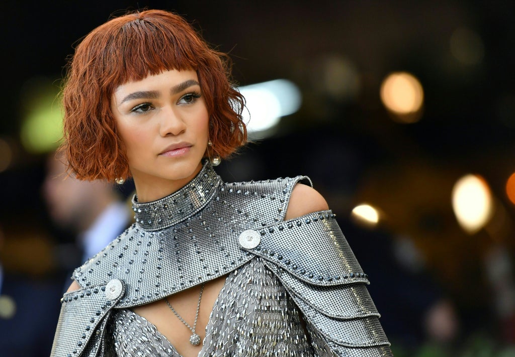 Zendaya wears a futuristic, armored silver ensemble with a structured shoulder piece and sparkling mesh detail, paired with a short auburn bob and blunt bangs. Her metallic look is complemented by delicate jewelry and radiant makeup.