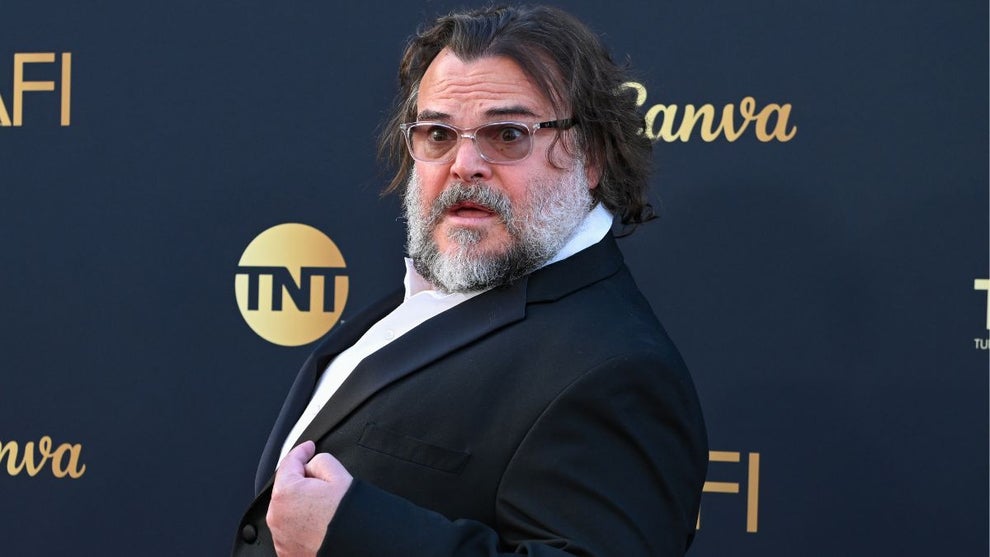 Jack Black strikes a surprised and playful pose on the red carpet while pointing at himself. He wears clear-framed glasses and a white shirt, with his signature bushy beard and tousled hair.