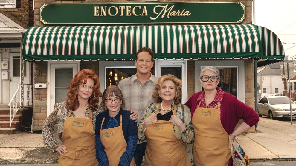 Vince Vaughn stands smiling behind four older women wearing aprons in front of Enoteca Maria, a real-life Staten Island restaurant known for its rotating lineup of grandmothers cooking international cuisine. The women, each with a distinct personality, stand proudly in front of the shop's green-and-white striped awning under the gold-lettered sign.