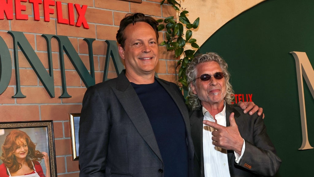 Vince Vaughn poses with Joe Scaravella at the Netflix premiere of Nonna’s. Vaughn, in a navy shirt and charcoal blazer, smiles with his arm around Scaravella, who wears a gray suit, white shirt, and sunglasses while pointing playfully at the camera. A framed still image from the film and the Netflix logo appear in the background.