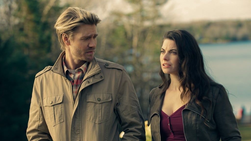 A man and woman stand outdoors near a lake, engaged in a serious conversation. The man, wearing a tan jacket over a plaid shirt, looks at the woman with concern. She wears a dark leather jacket over a burgundy top, her long dark hair blowing slightly in the breeze. There are scenic trees and a waterfront in the background.