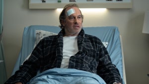A middle-aged man with graying hair and a beard sits upright in a hospital bed, wearing a black plaid robe over a white shirt. A large bandage is taped to his forehead, and his expression is dazed and concerned as he looks off to the side. The clinical setting and medical equipment in the background emphasize the seriousness of the moment.