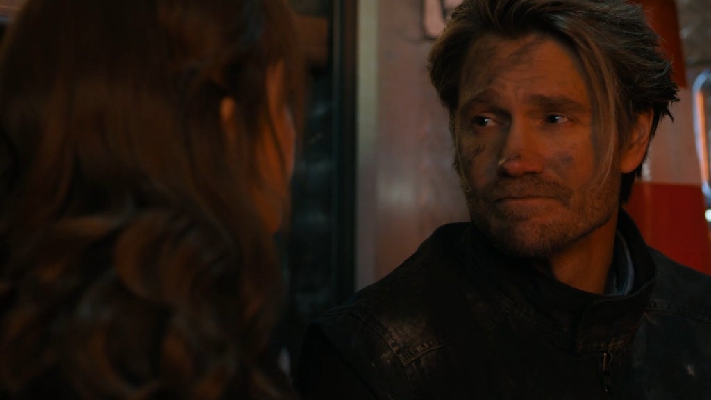 A man with dirt and soot on his face, wearing a black jacket, looks emotionally at a woman whose face is mostly out of frame. His expression is somber and reflective, suggesting the aftermath of a traumatic event. The warm lighting and background hint at an emergency scene.