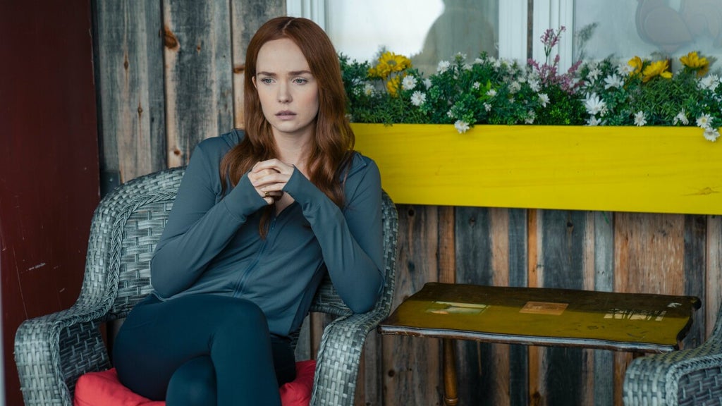 A red-haired woman sits pensively on a wicker chair outside a rustic wooden building, her hands clasped in front of her as she stares off thoughtfully. She wears a dark zip-up top and leggings, with a bright yellow flower box full of blooms behind her.