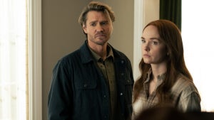 A man and woman stand side by side in a softly lit room, both wearing serious, concerned expressions. The man wears a dark jacket over an olive shirt, while the woman, with long auburn hair, wears a light plaid button-down. They look ahead with intense, focused expressions.