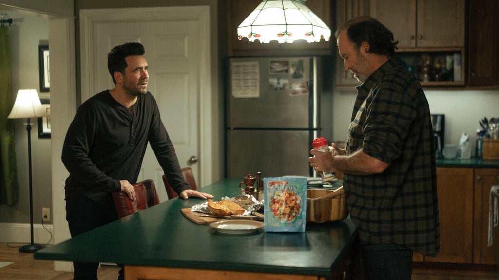 Two men talk in a warmly lit kitchen. One, dressed in a dark long-sleeve shirt, leans against a counter with a serious expression, while the other, in a plaid shirt, prepares food beside a salad bag and garlic bread. 