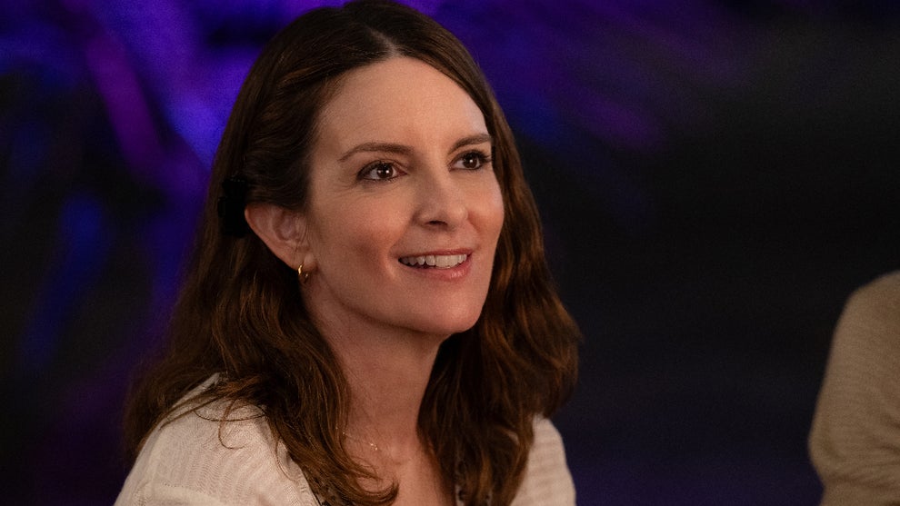A woman with brown hair in a white flowing top smiles at someone out of frame. There is blue light behind her.