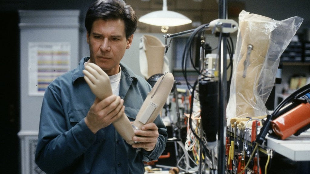 Harrison Ford, as Dr. Richard Kimble in The Fugitive, examines a prosthetic arm in a cluttered lab filled with wires, tools, and mannequin limbs. He wears a serious expression and a blue work shirt as he investigates a key clue in the suspenseful thriller.