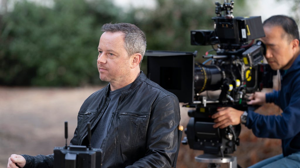 A man with short greying hair wears a leather jacket and sits on a TV set. There is a man holding a big camera and lens behind him. 