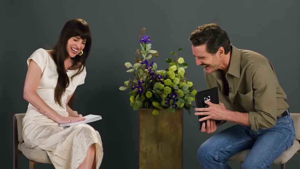 Dakota Johnson and Pedro Pascal bend over laughing in an interview setting, both holding cue cards. She wears a white dress. He wears a neutral shirt and blue jeans.