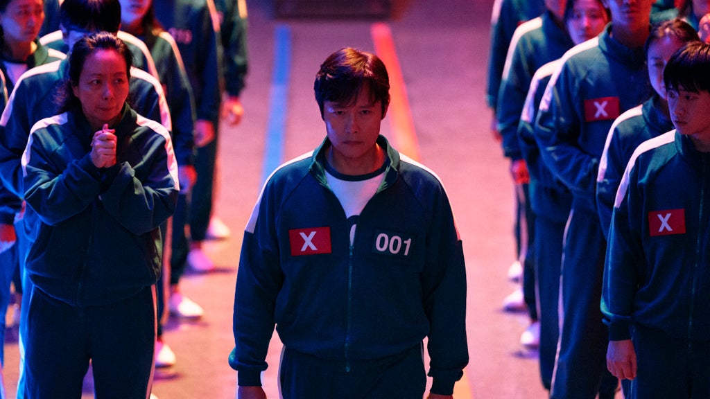 A man wearing a green tracksuit labeled “001” and marked with a red “X” walks forward with a stern, determined expression. Behind him, rows of other contestants in identical tracksuits also wear red “X” patches