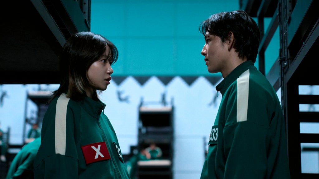 A young woman and man in green tracksuits stand face-to-face in a tense exchange under the dim lighting of a dormitory filled with black bunk beds.