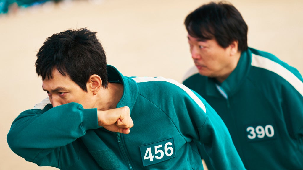 Two men in teal tracksuits with white stripes, numbered 456 and 390, appear tense and mid-motion during a high-stakes game. The man in front, number 456, covers his face with his arm in a defensive gesture, while the man behind him looks forward intently.