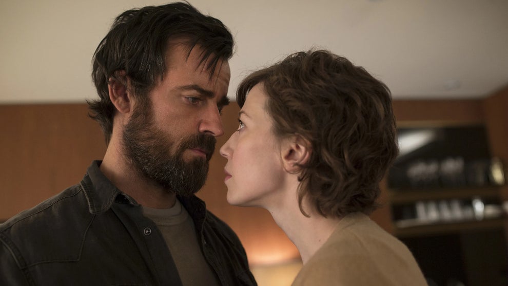 A man and woman stand face to face in intense eye contact, their expressions serious and emotional. The man has a rugged beard and dark hair, while the woman has short, wavy hair.