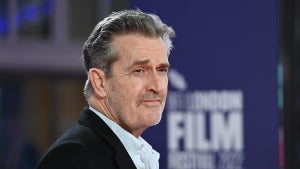 Rupert Everett wears a suit on a red carpet, shot from the shoulders up, in front of a purple backdrop that says BFI LONDON FILM FESTIVAL 2022
