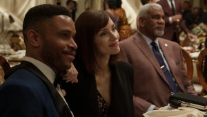 A woman sits at a table smiling, posing with a man to her right. Another man sits on her left