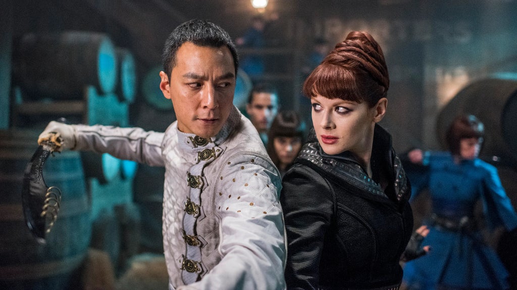 A man and a woman stand back-to-back, poised for a fight with people surrounding them