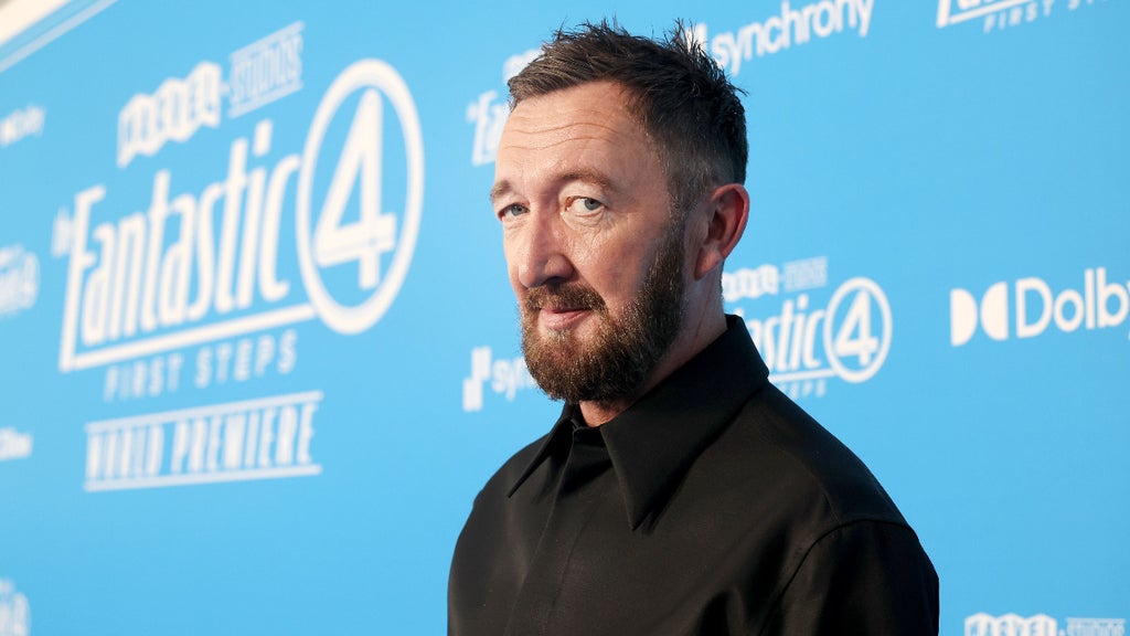 Ralph Ineson smiles at the camera on the "Fantastic Four: First Steps" red carpet with a blue logo backdrop behind him