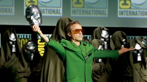 Robert Downey Jr. stands on stage at Comic-Con in a Victor Doom costume, hold his mask toward the sky in a triumphant gesture